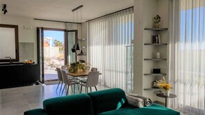A modern living room with a green sofa, a dining table with chairs, and a kitchen area visible through an open door. - luxury holiday home in Lo Stagnone I Palm Kite Paradise I Casa A (Marsala)