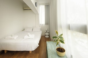 A bedroom with a bed, a bedside table with a potted plant, and a window with sheer curtains.