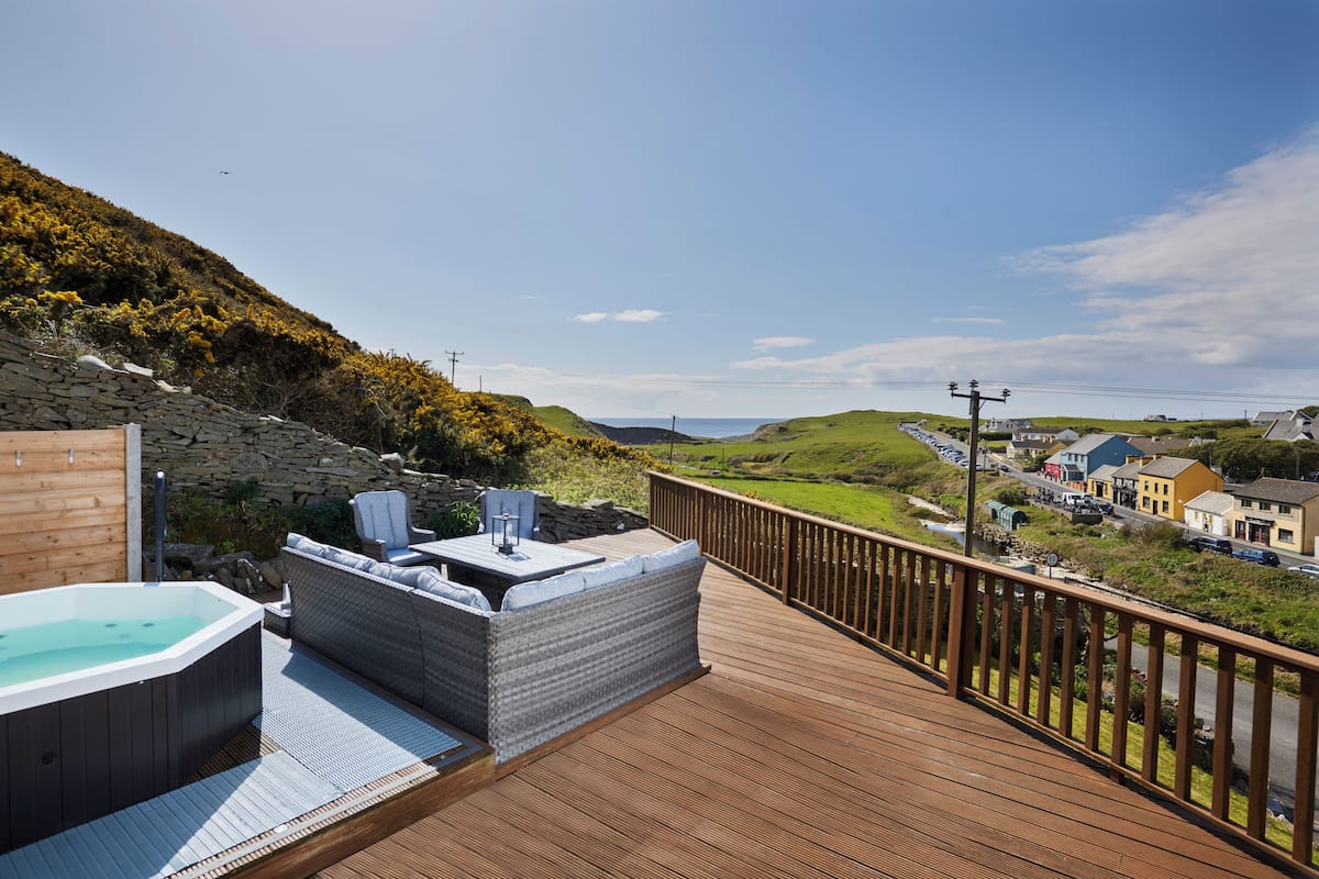 Eine Holzterrasse mit Whirlpool und Sitzgelegenheit mit Blick auf eine malerische Landschaft aus Häusern und Hügeln.
