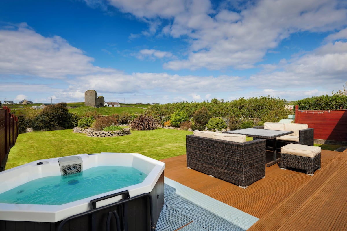 Ein Whirlpool auf einer Holzterrasse mit Korbmöbeln, einem Rasen und einer Steinstruktur im Hintergrund.