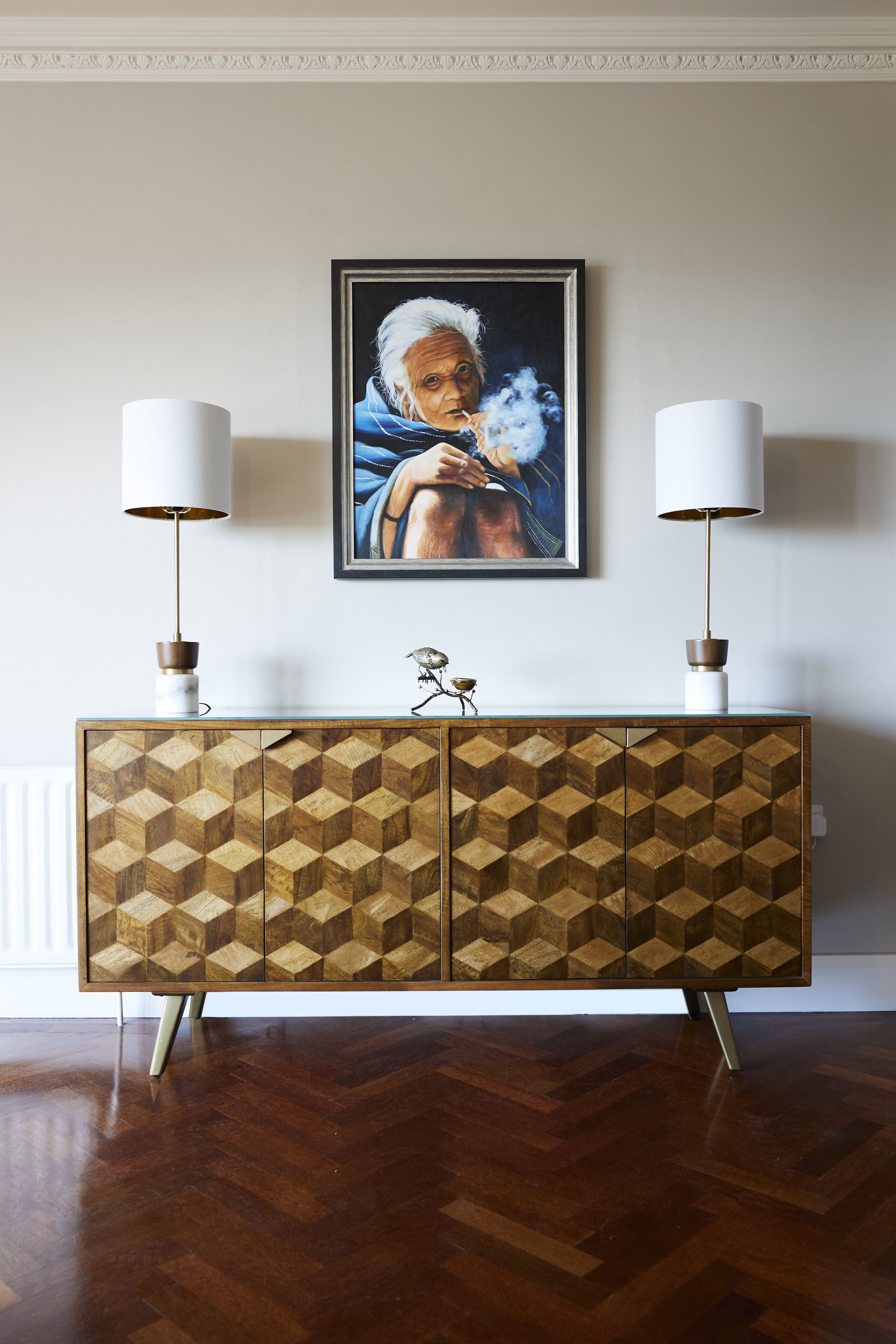 A wooden cabinet with geometric patterns, flanked by two white lamps with brown bases, and a framed painting above.