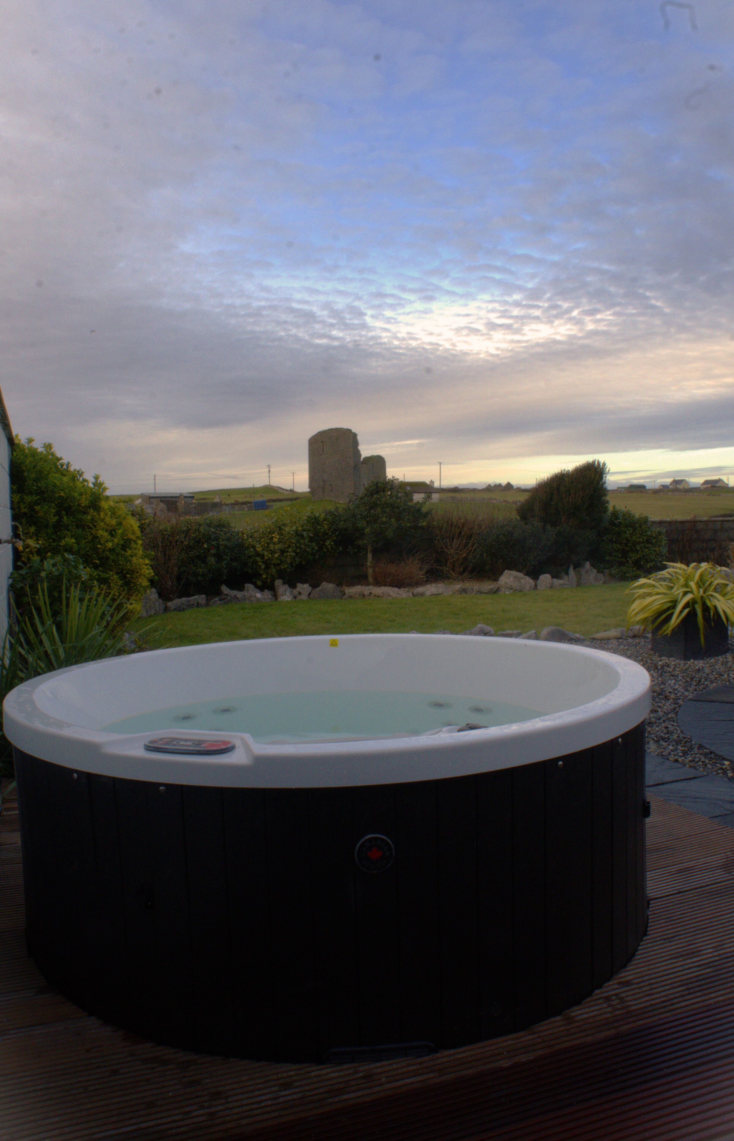 A hot tub on a wooden deck with a view of a grassy area and a stone structure in the background.
