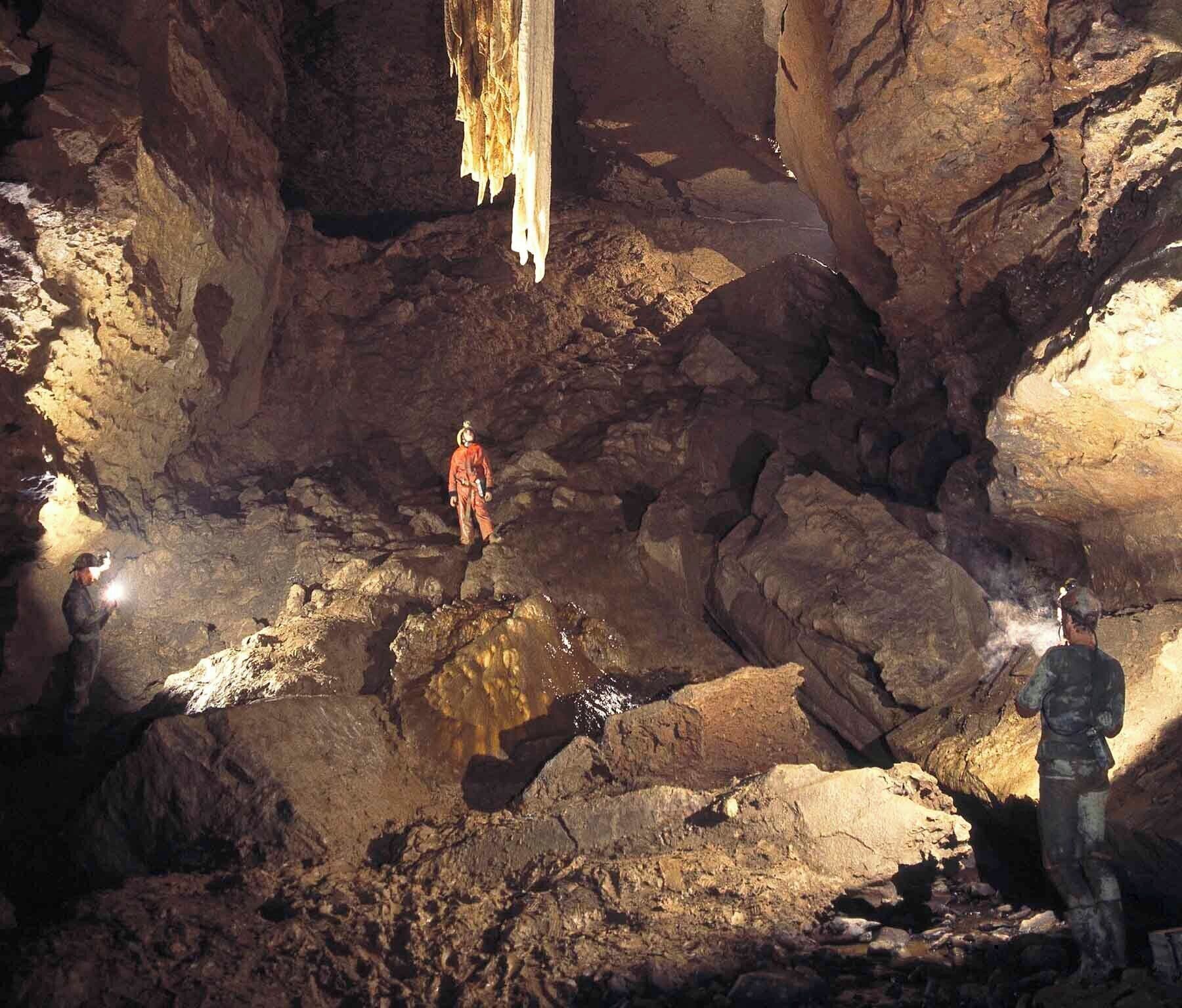 Una cueva con estalactitas y estalagmitas, terreno rocoso y dos personas explorando.