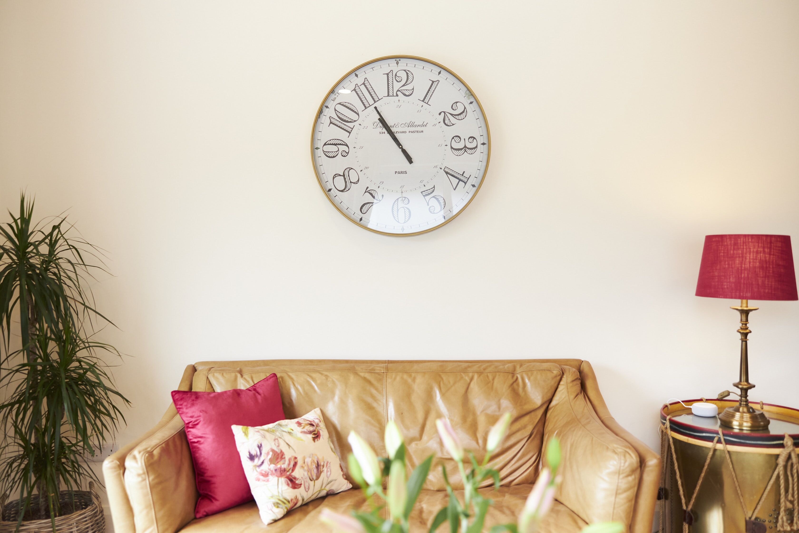 A room with a beige leather sofa, a round wall clock, a potted plant, and a lamp.