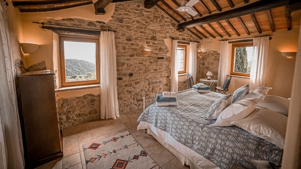 Ein Schlafzimmer mit Steinwand, Holzboden, einem Bett mit weißblauer Bettwäsche, einem kleinen Tisch mit Lampe und Blick ins Freie.