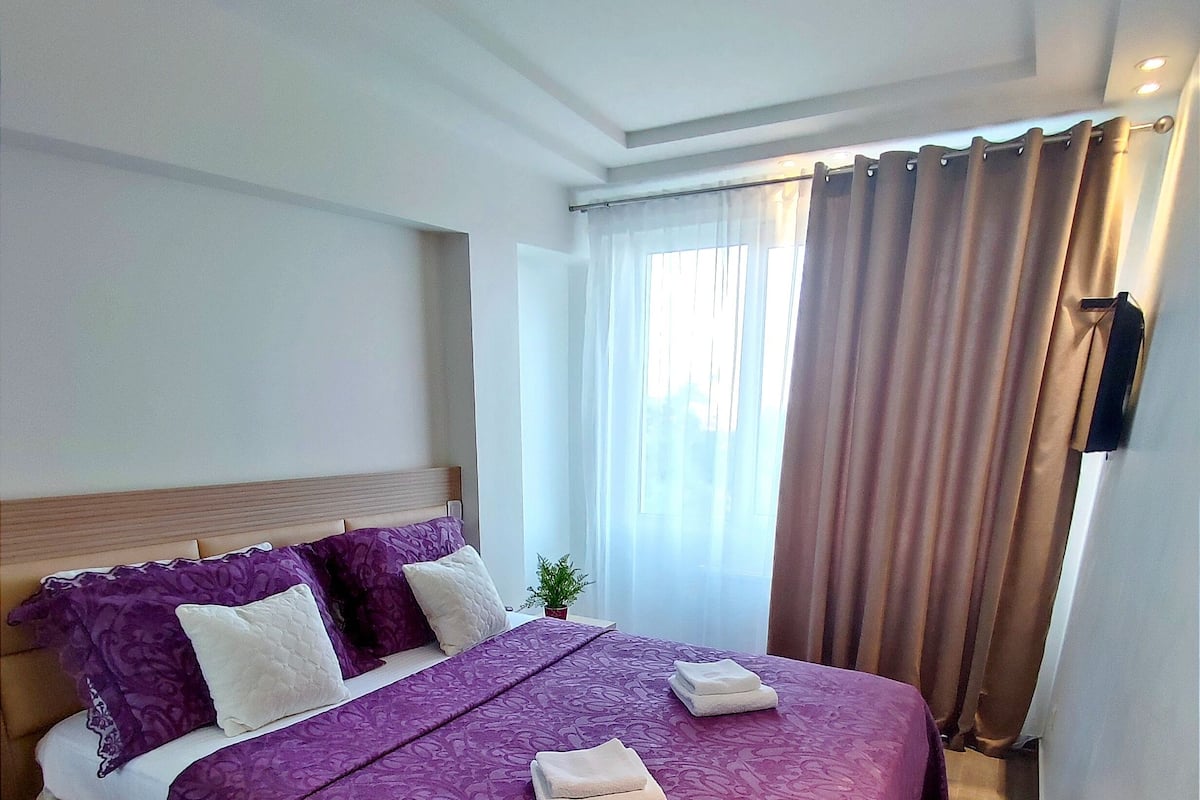 Een hotelkamer met een groot bed, een houten hoofdeinde, een kleine plant in een pot en een raam met gordijnen.