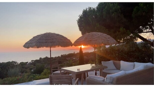 A sunset view with two thatched umbrellas, a wicker sofa, and a table on an outdoor terrace.
