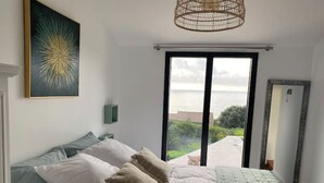 A bedroom with a bed, pillows, a bedside table, a wall-mounted light, a large window with a view of the sea, and a door.