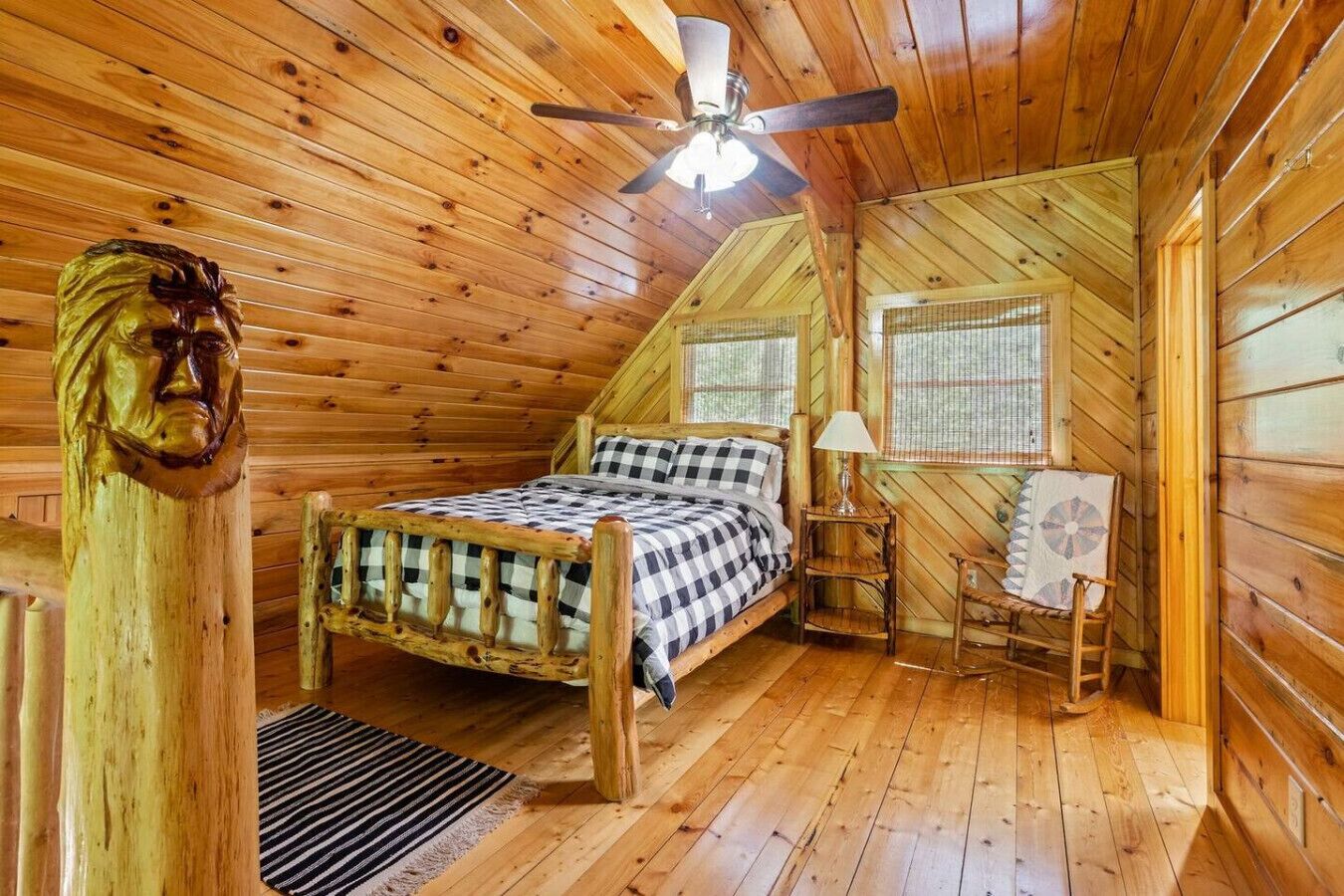 A cozy wooden cabin bedroom with a bed, a small table, a chair, and a ceiling fan.