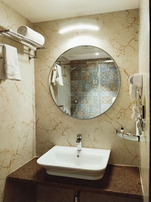 A bathroom with a marble vanity, a round mirror, a towel rack with towels, a shower with a tiled wall, and a hairdryer mounted on the wall.