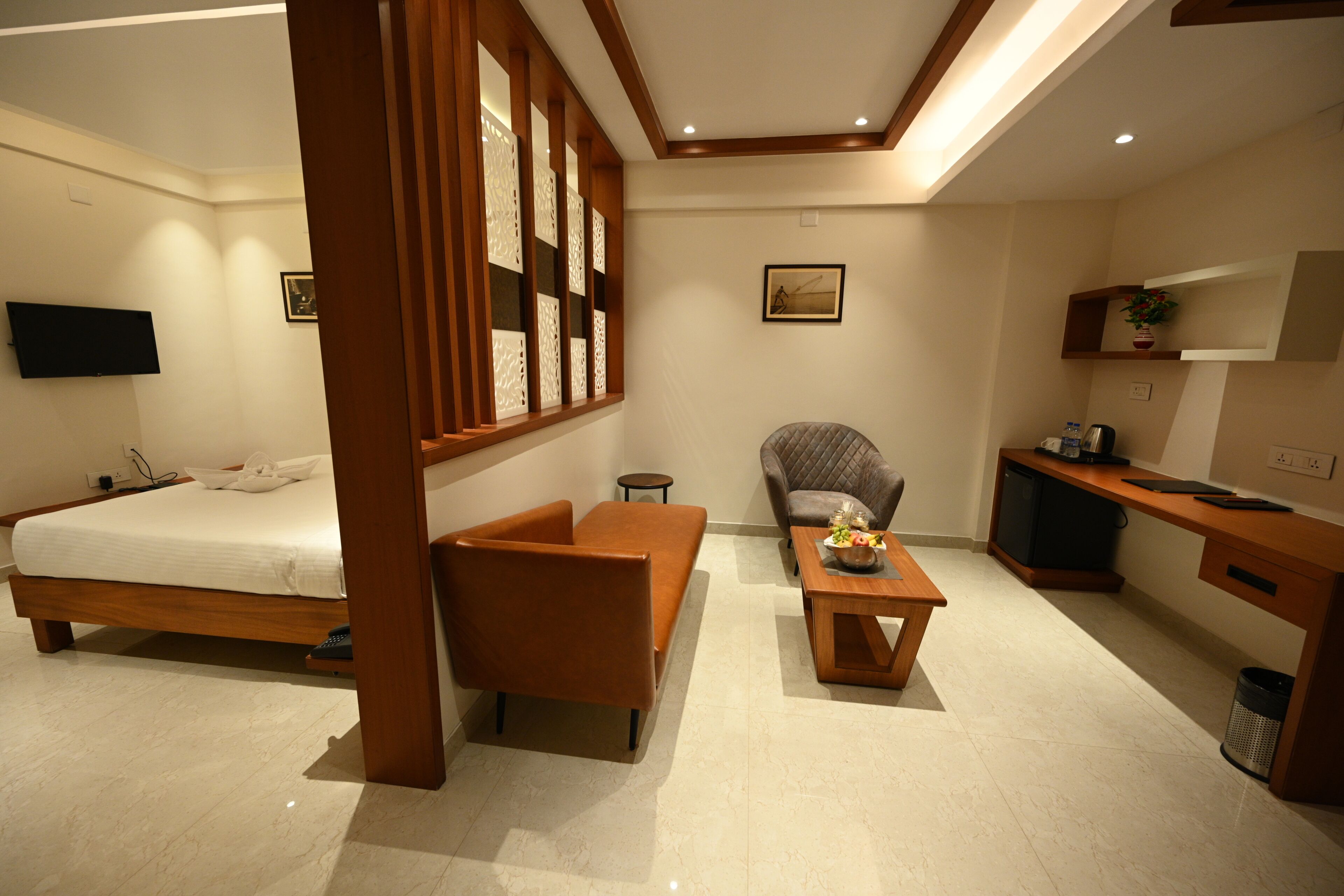 A modern hotel room with a bed, a sofa, a small table with a vase, a chair, a desk, and a television on the wall.
