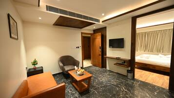 Habitación de hotel con cama, televisión, zona de estar con una silla y una mesa, y una planta pequeña en la mesita de noche.
