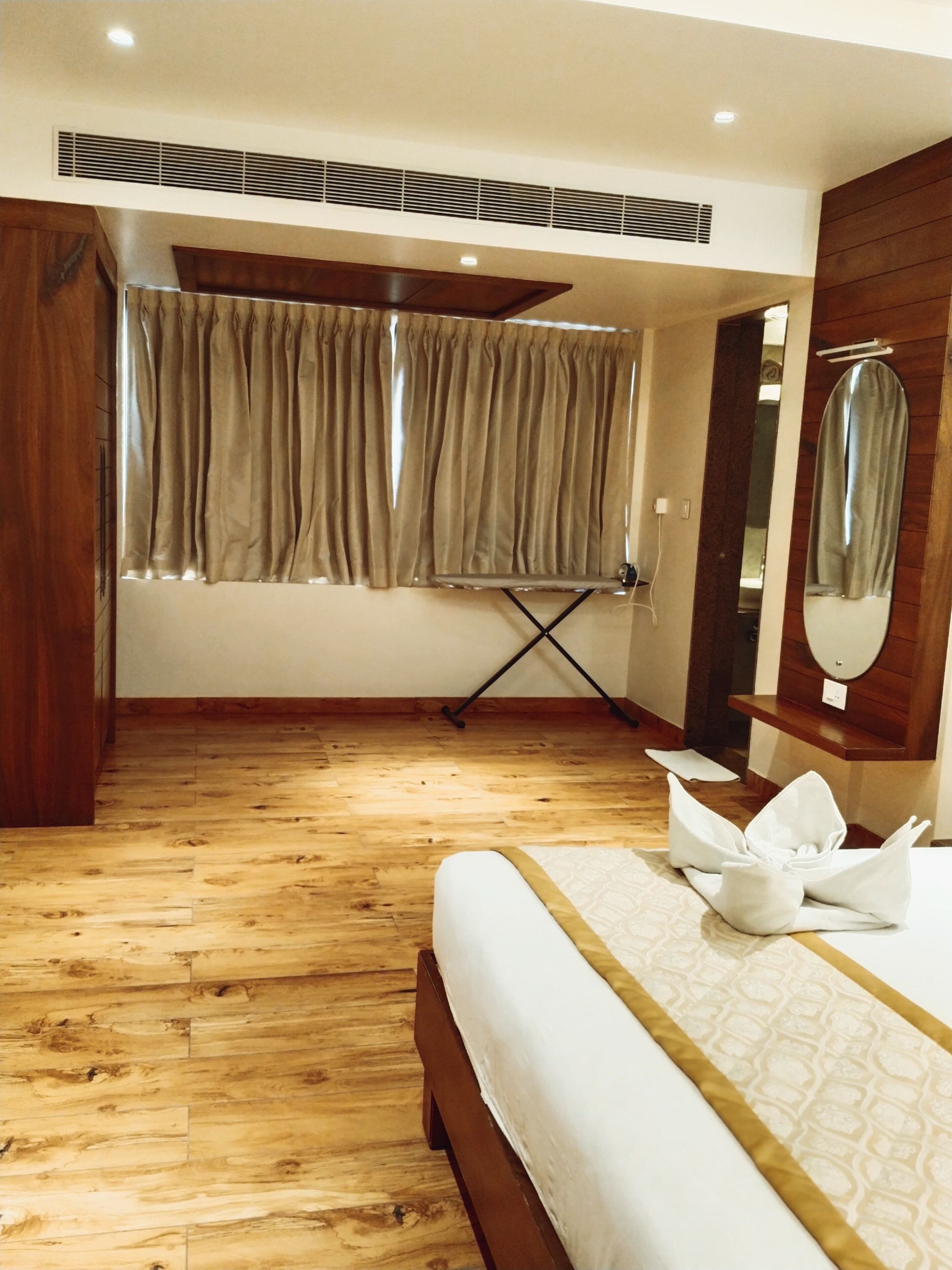 A hotel room with a wooden floor, a bed with white bedding, a window with beige curtains, a mirror, and a small table with a towel.