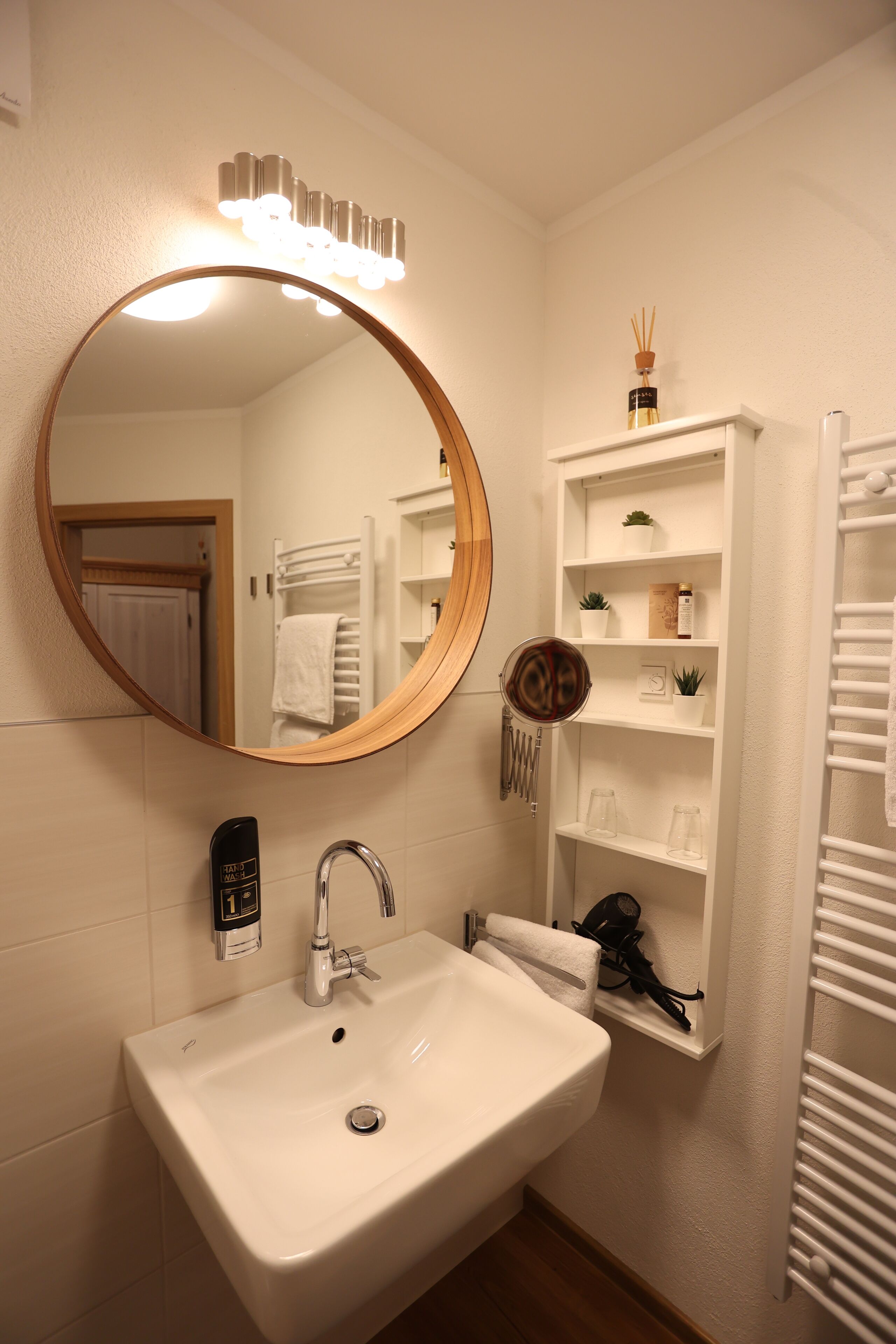 A bathroom with a round mirror, a white sink, a towel rack, and a shelf with toiletries.