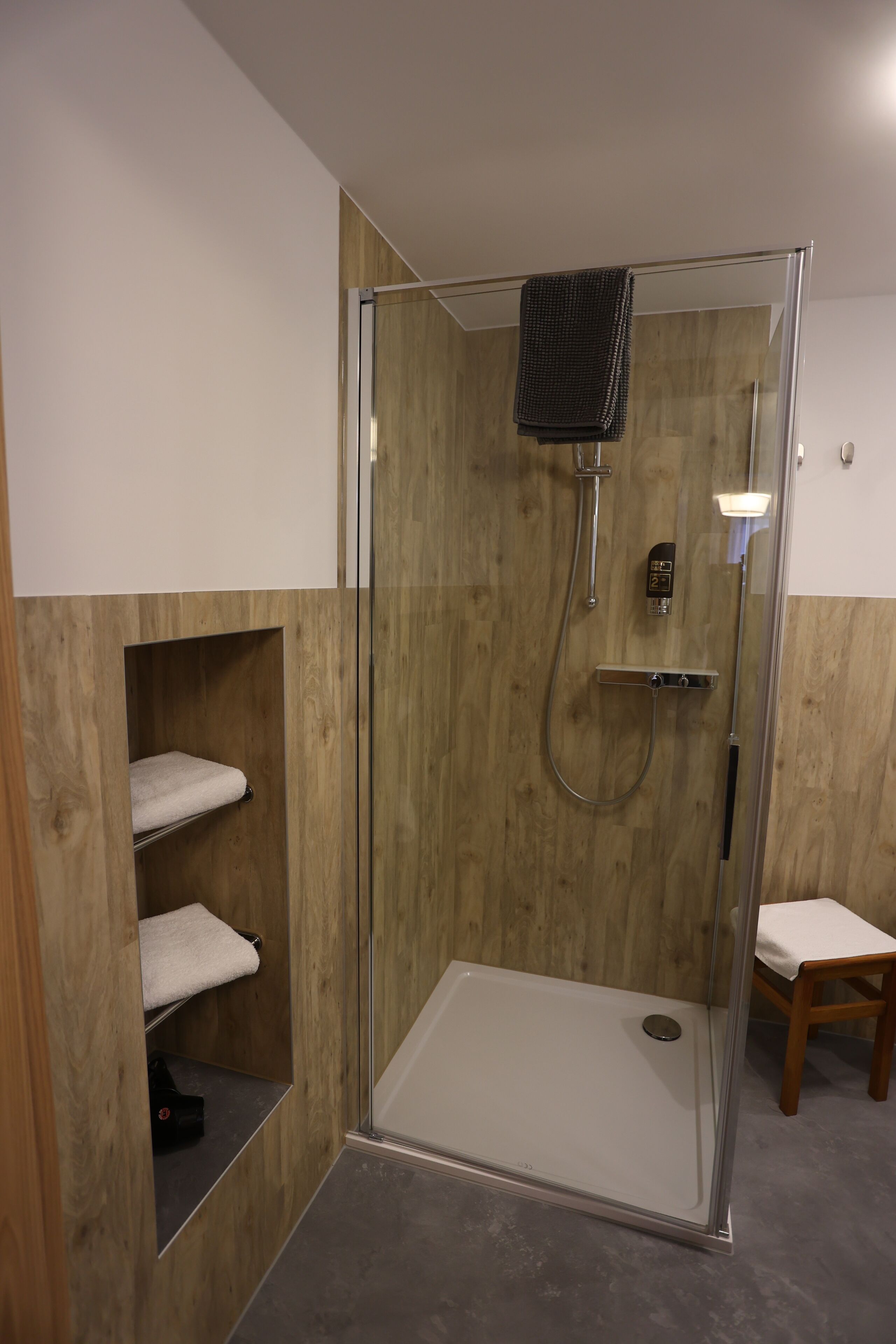 A modern bathroom with a glass-enclosed shower, wooden paneling, a towel rack, and a wooden chair.