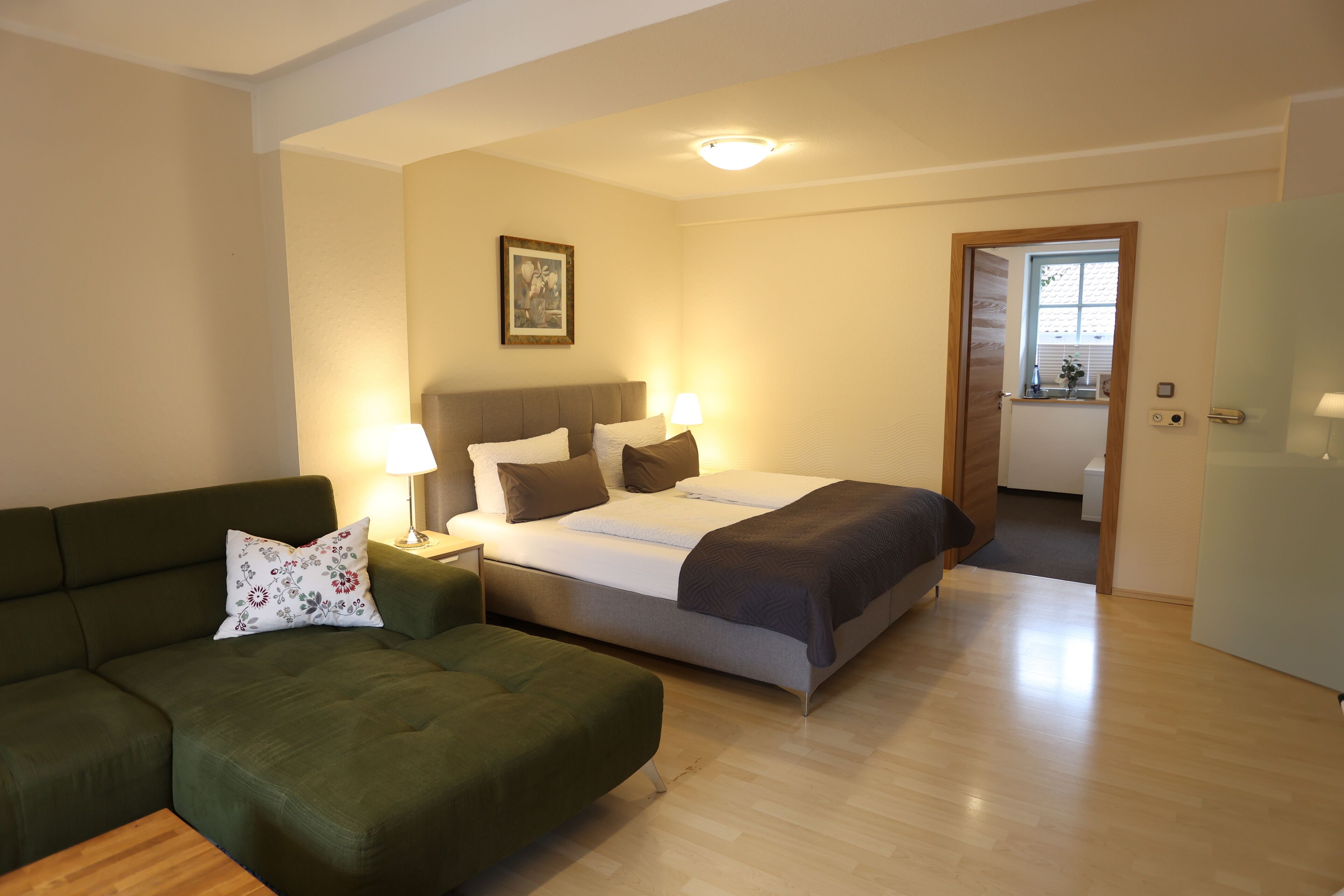 A hotel room with a bed, a green sofa, a nightstand with lamps, and a door leading to a bathroom.