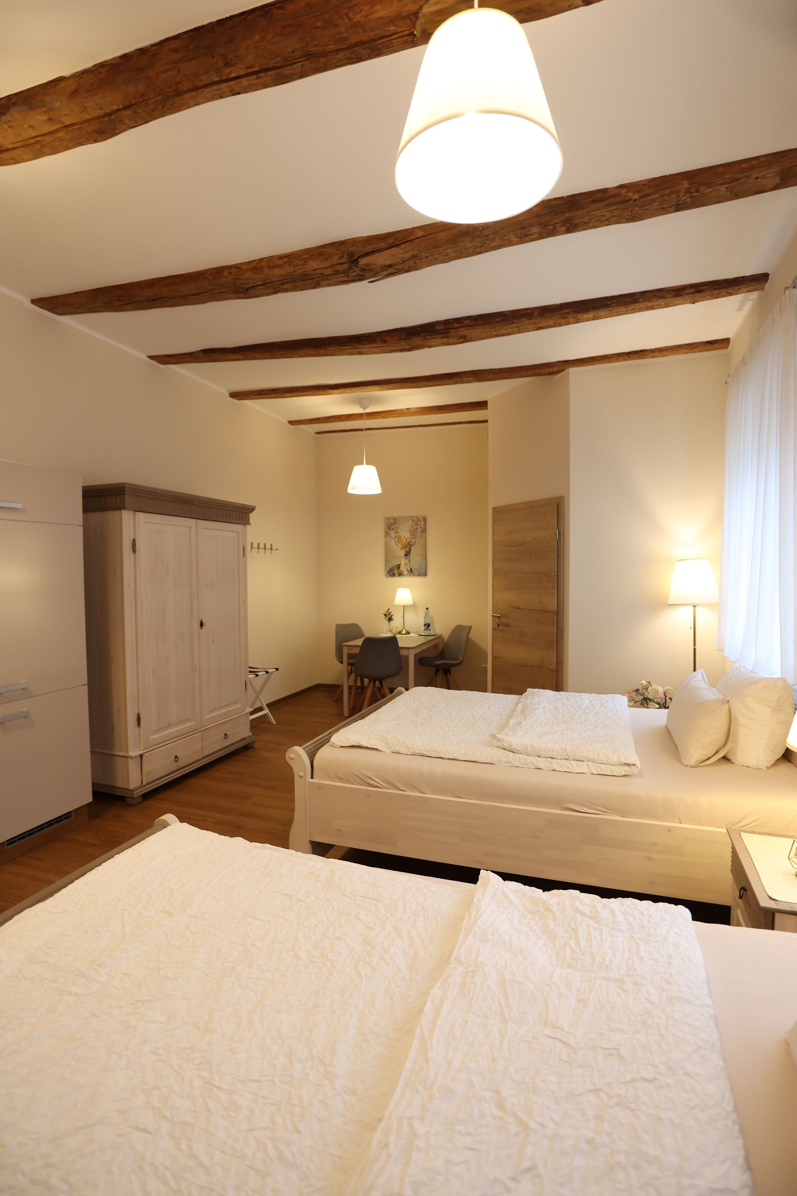 A hotel room with two beds, wooden beams on the ceiling, a desk with a chair, and a wardrobe.