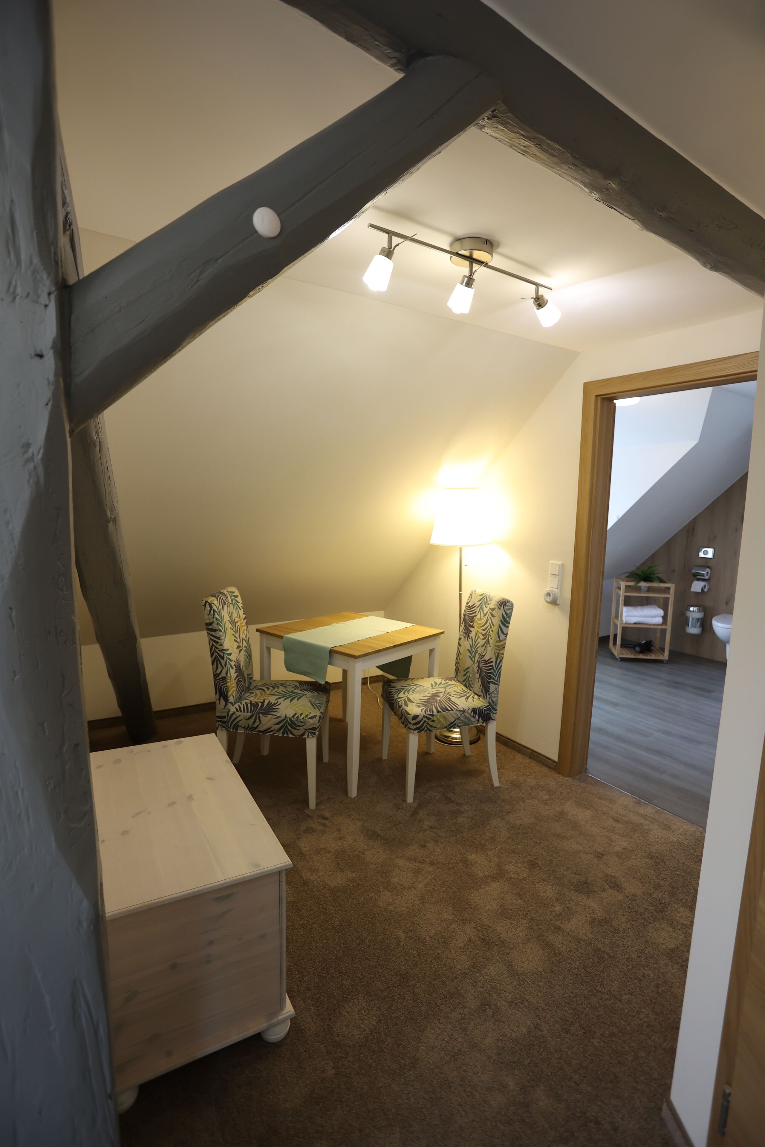 Una habitación pequeña con una mesa, dos sillas, una lámpara y un armario de madera.