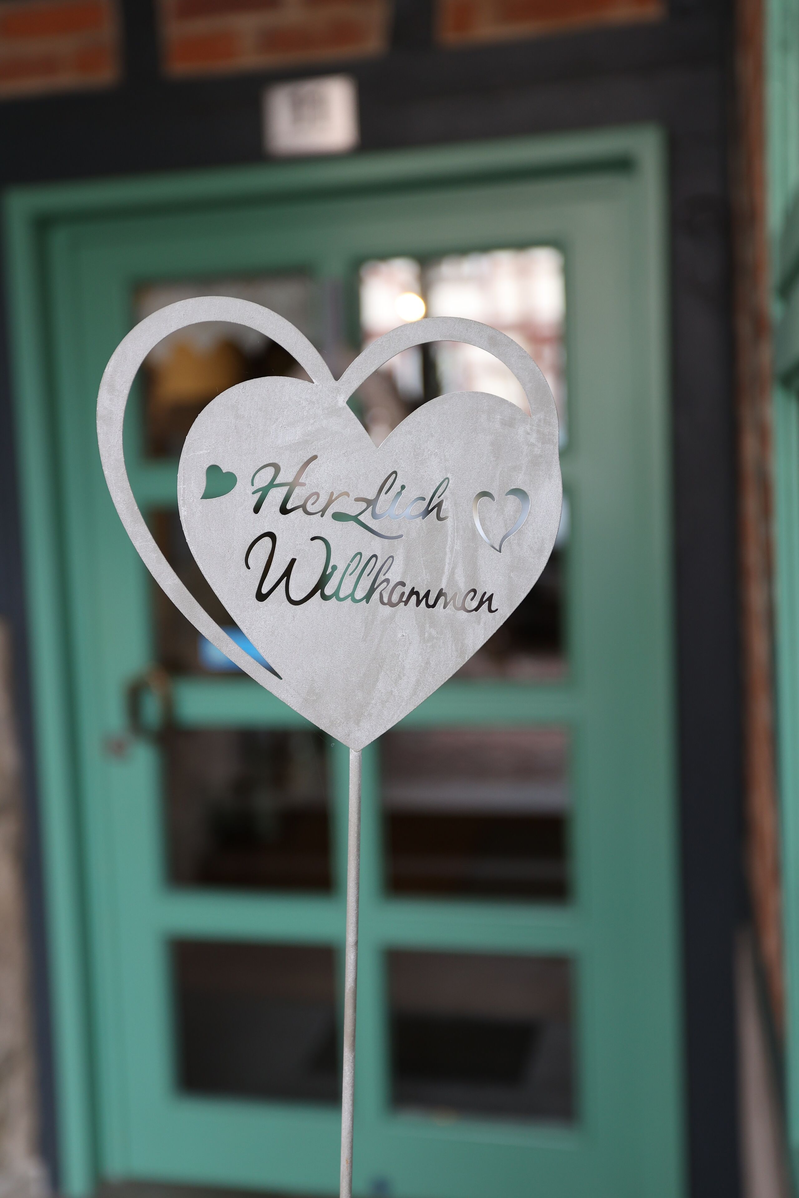 A heart-shaped sign with the words "Herzlich Willkommen" on a stick, in front of a teal door with windows.