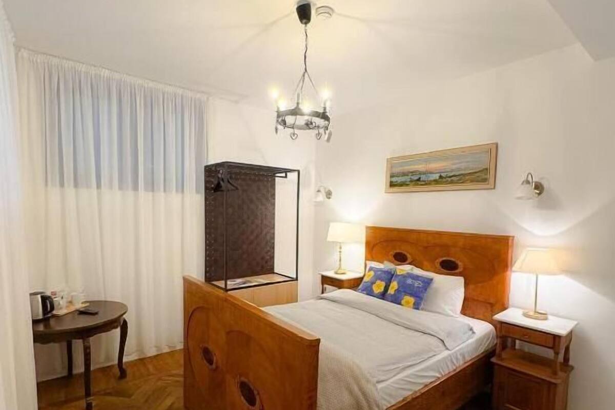 A bedroom with a wooden bed, a small bedside table, a nightstand, a chandelier, a painting on the wall, and a window with curtains.