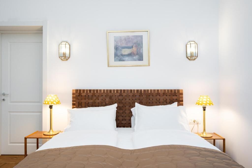 Un letto rifatto con cura, con una testiera in legno, due lampade sui comodini e un quadro appeso sopra il letto.
