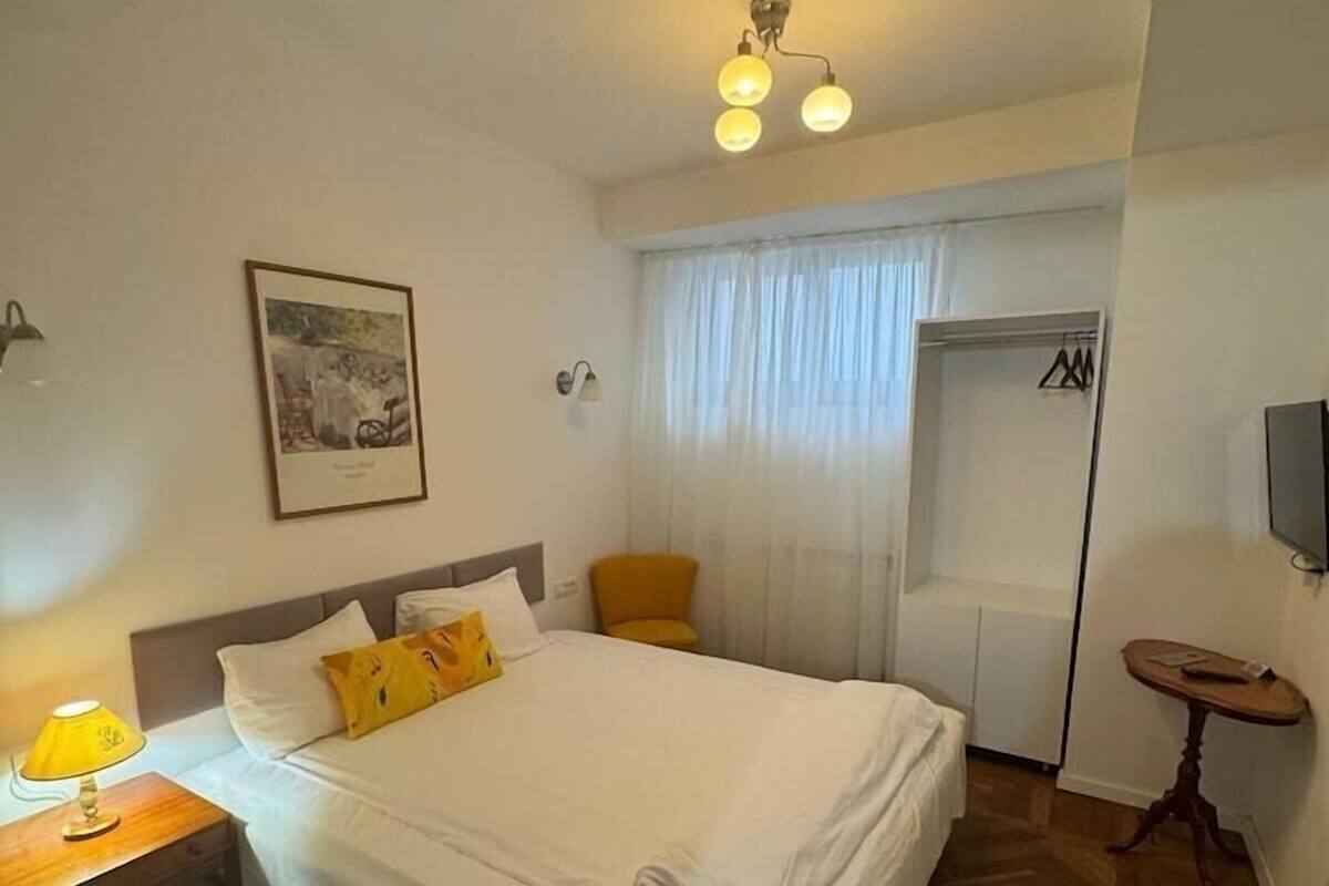 A hotel room with a bed, a desk with a lamp, a chair, a wardrobe, and a television.