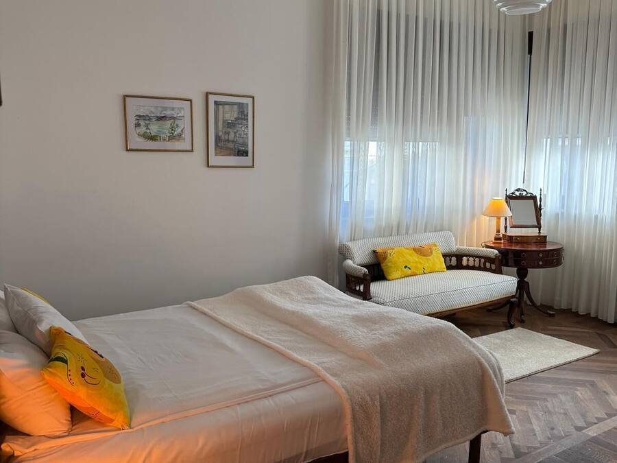 A bedroom with two beds, a bedside table with a lamp, a chair, and a small table with a lamp.