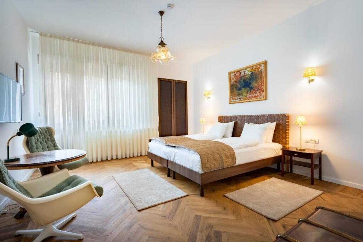 A hotel room with a bed, a chair, a table, and a painting on the wall.