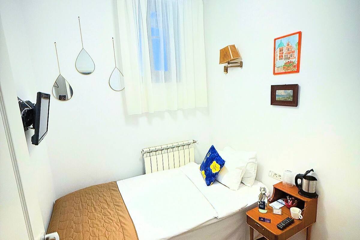 A small, tidy bedroom with a single bed, a bedside table with a kettle and a cup, a radiator, and a window with curtains.