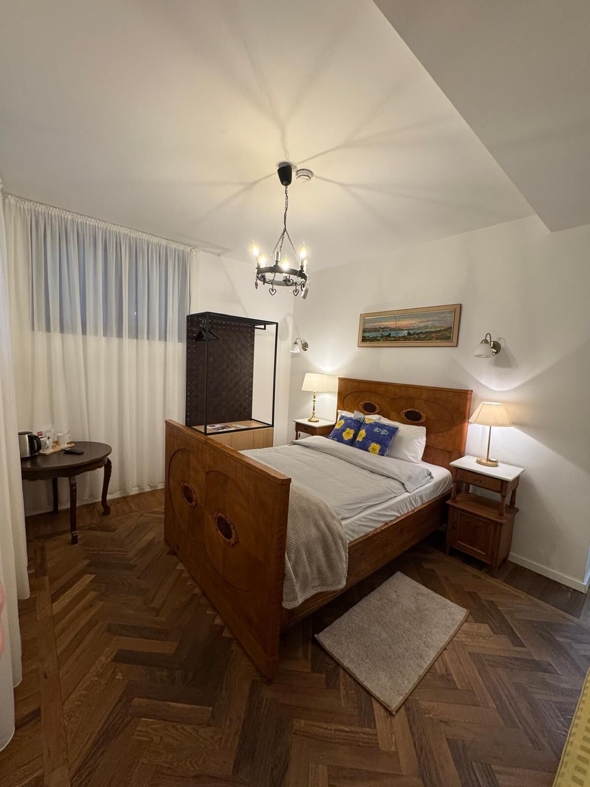 Une chambre à coucher avec un lit en bois, une table de chevet, une lampe de chevet, un lustre, un tableau au mur et une vue sur l’extérieur par la fenêtre.