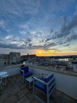 A rooftop terrace with a view of the sunset over a cityscape, featuring a table and two blue cushioned chairs. - San Pietro Otranto  (Otranto)