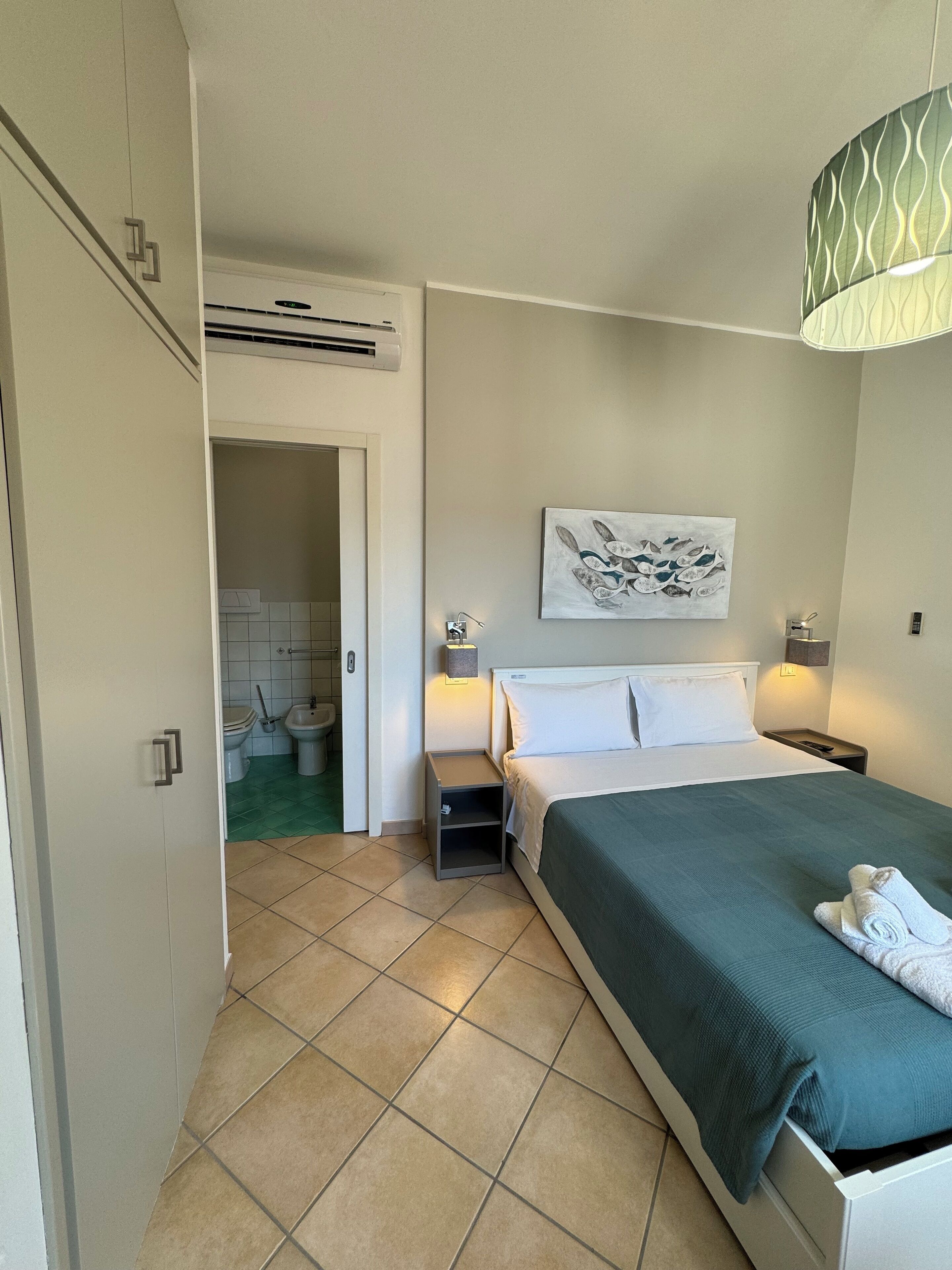 A hotel room with a bed, bedside tables, a wall-mounted artwork, and an air conditioning unit.