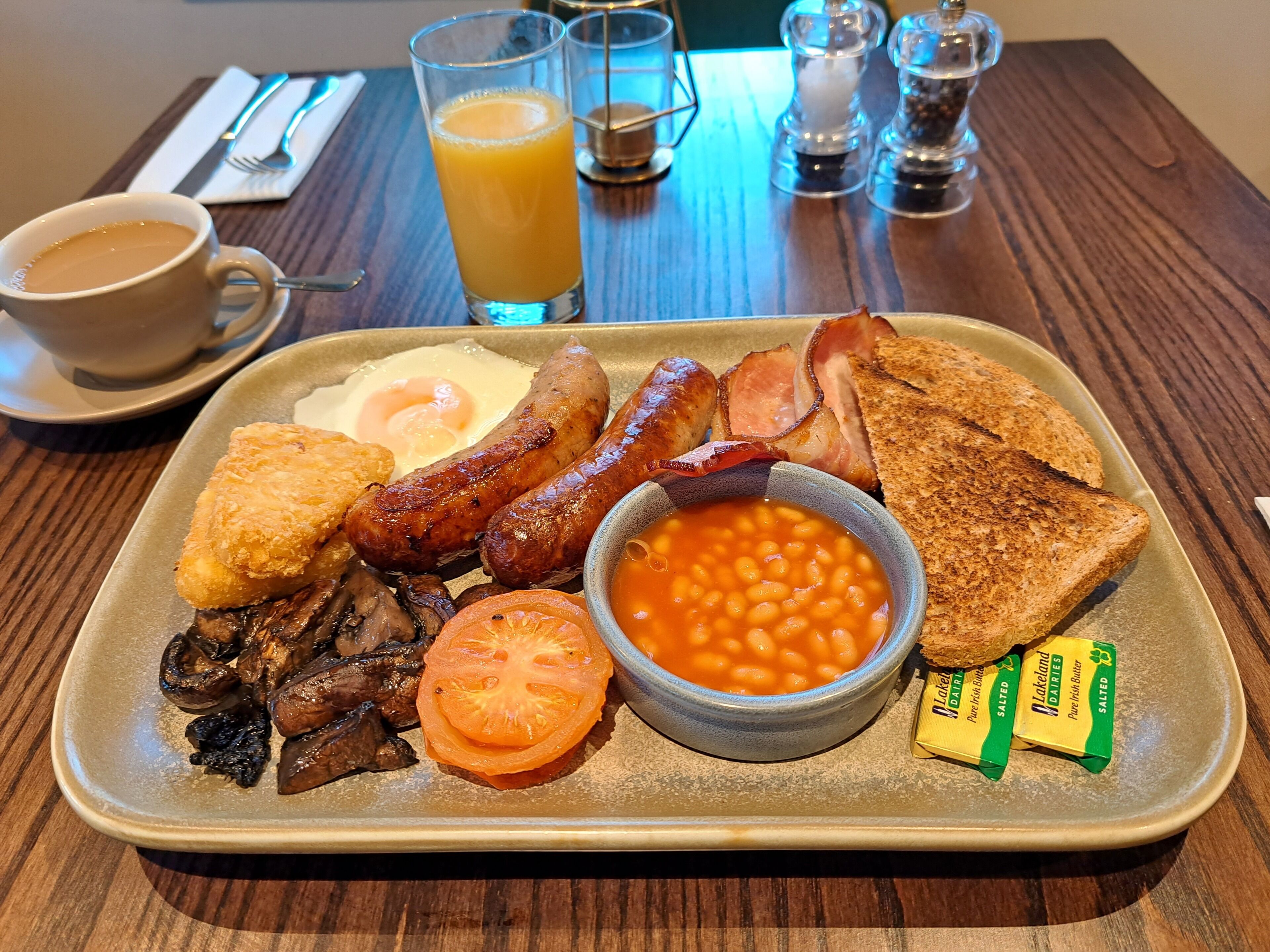 Un plateau de petit-déjeuner avec des œufs, des saucisses, du bacon, des toasts, des haricots, des champignons, une tranche de tomate, du beurre et une tasse de café.