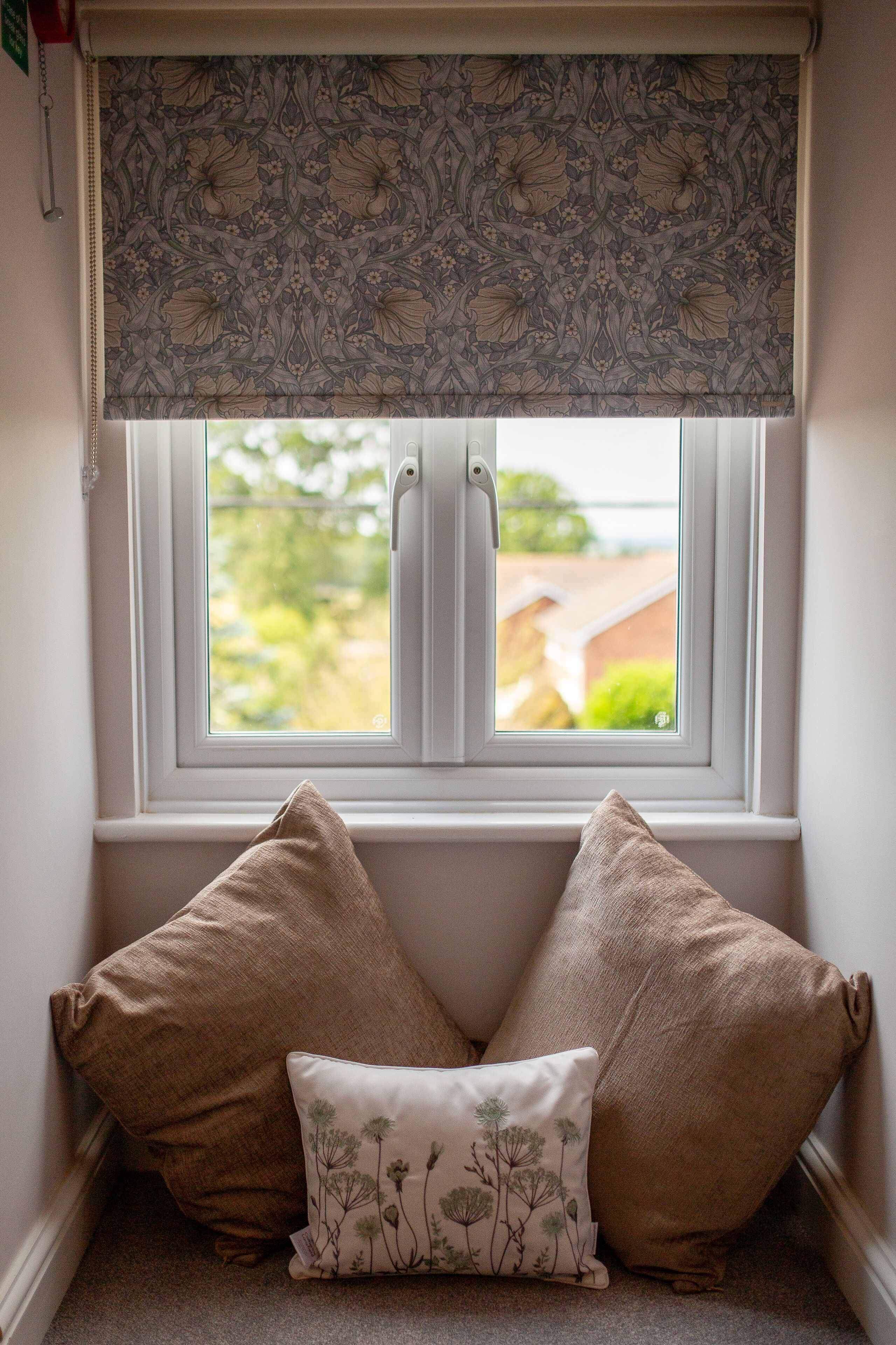 A corner window with a patterned roller blind, two beige pillows, and a small decorative pillow.