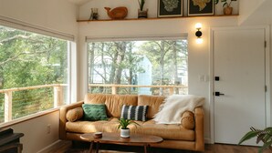 A cozy living room with a leather sofa, a coffee table, and a large window offering a view of trees. - The Weekender | Hot Tub| Neskowin Village (Neskowin)