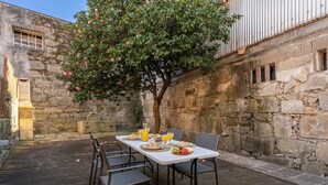 Zona pranzo all'aperto con parete in pietra, terrazza in legno e tavolo per due apparecchiato con piatti, bicchieri e una ciotola di frutta.