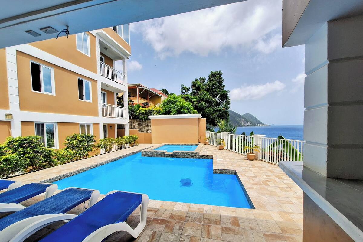 A swimming pool with lounge chairs, a view of the sea, and a building with balconies.
