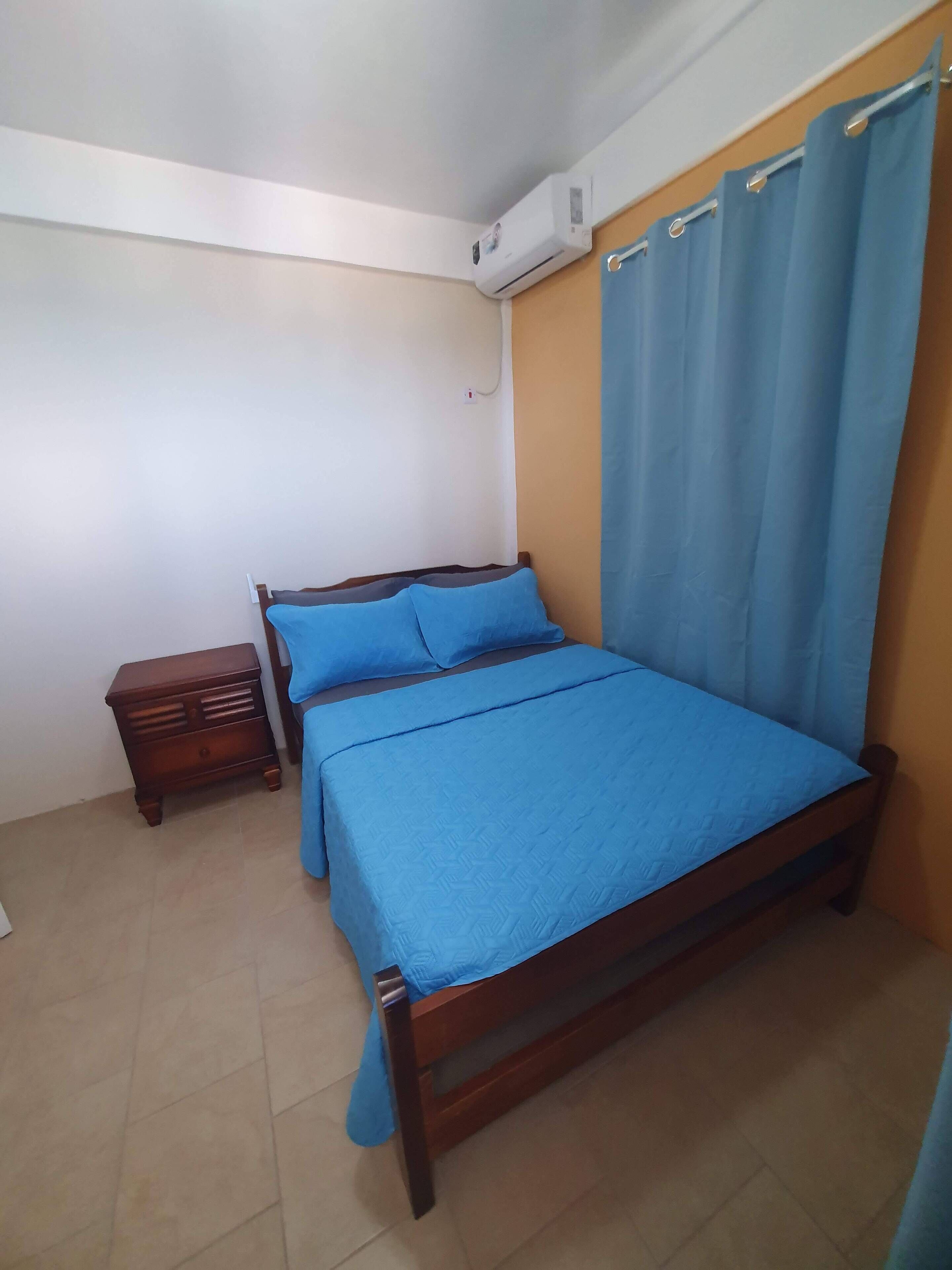 Un dormitorio con una cama de madera, ropa de cama azul, una mesita de noche y un equipo de aire acondicionado en la pared.