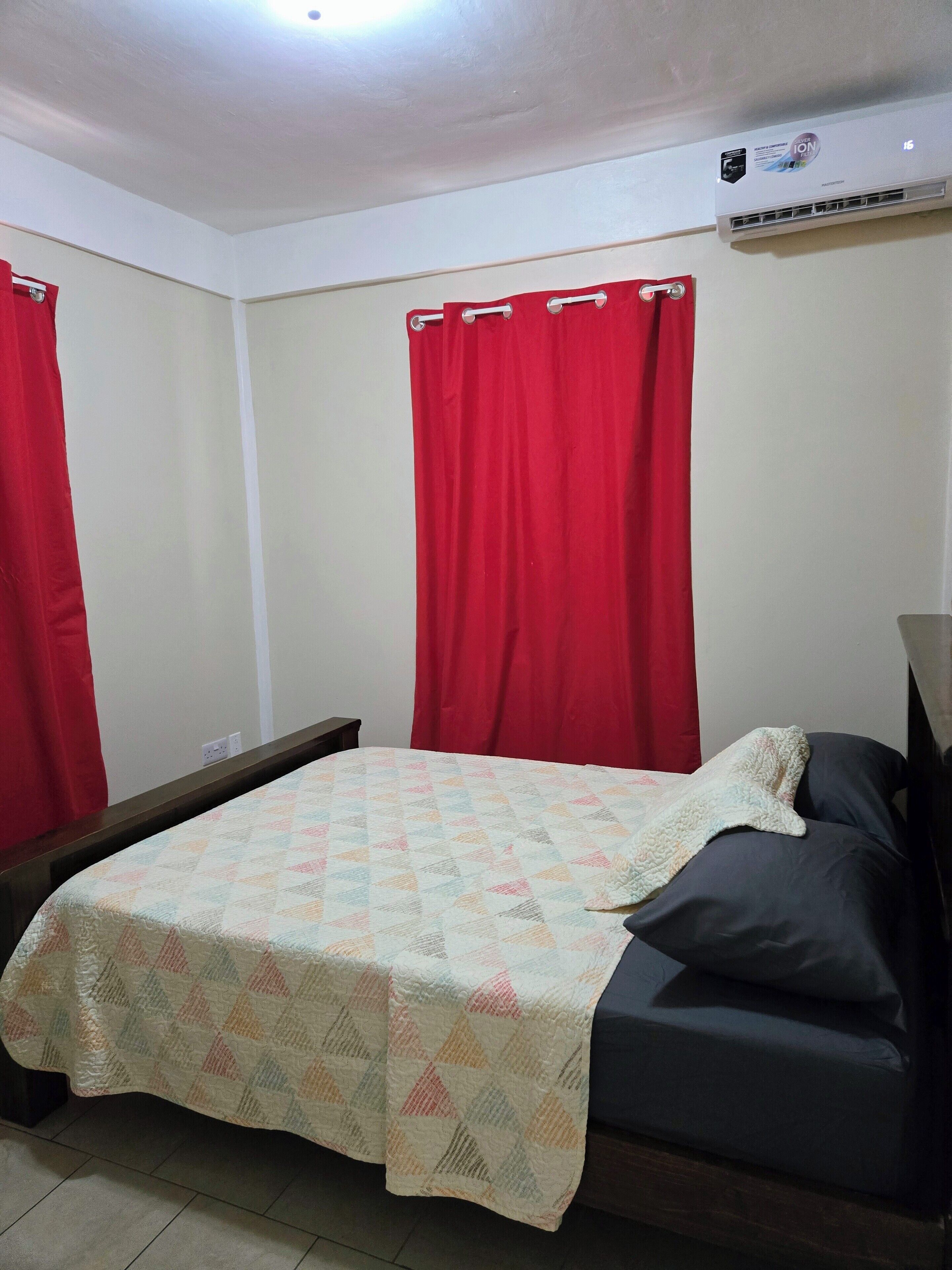 A room with a bed, red curtains, a white wall, and an air conditioning unit.