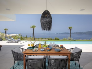 A dining area with a view of the sea, a pool, and lounge chairs.
