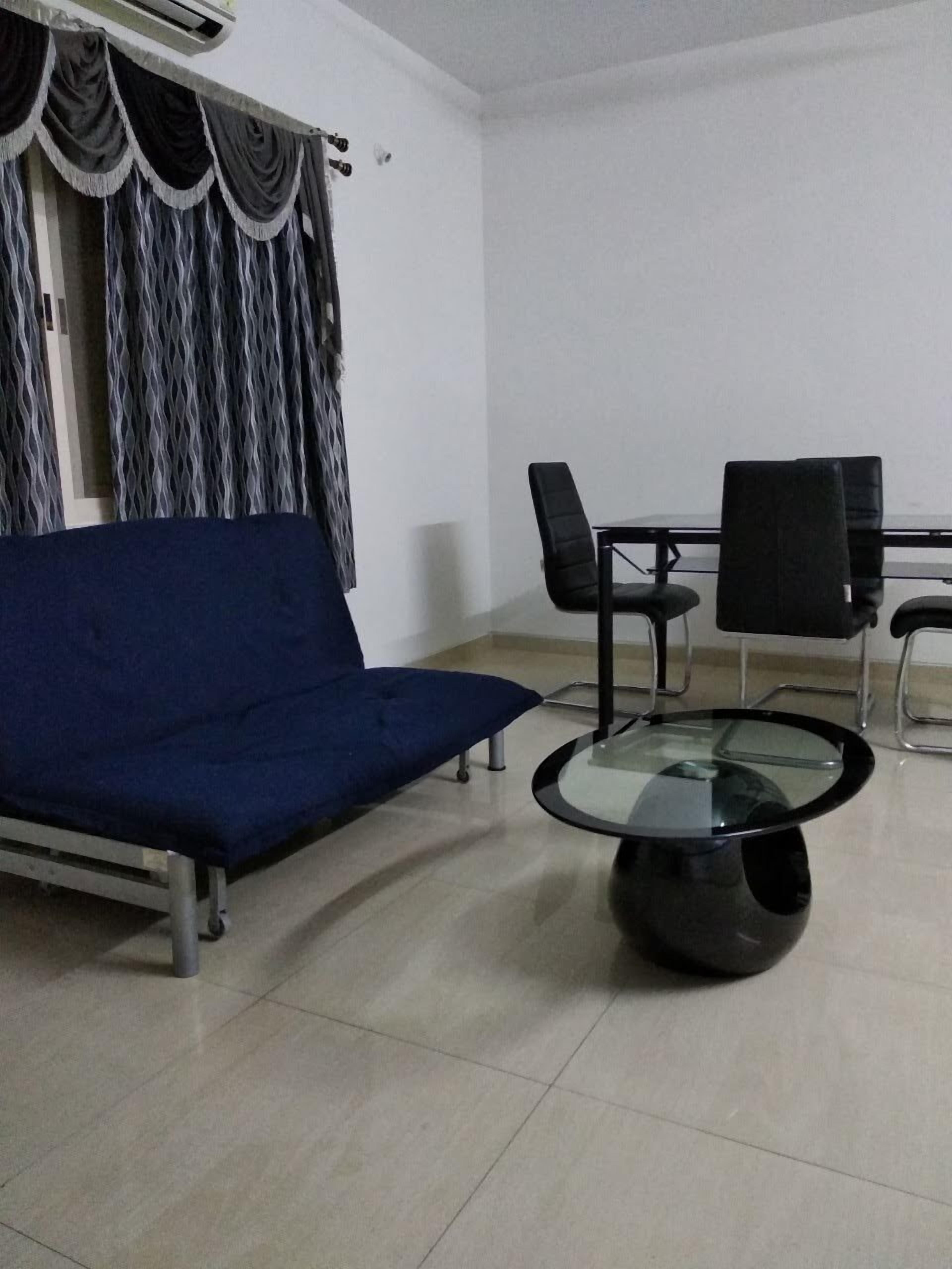 Quarto com um sofá azul, uma mesa com tampo de vidro e duas cadeiras pretas.