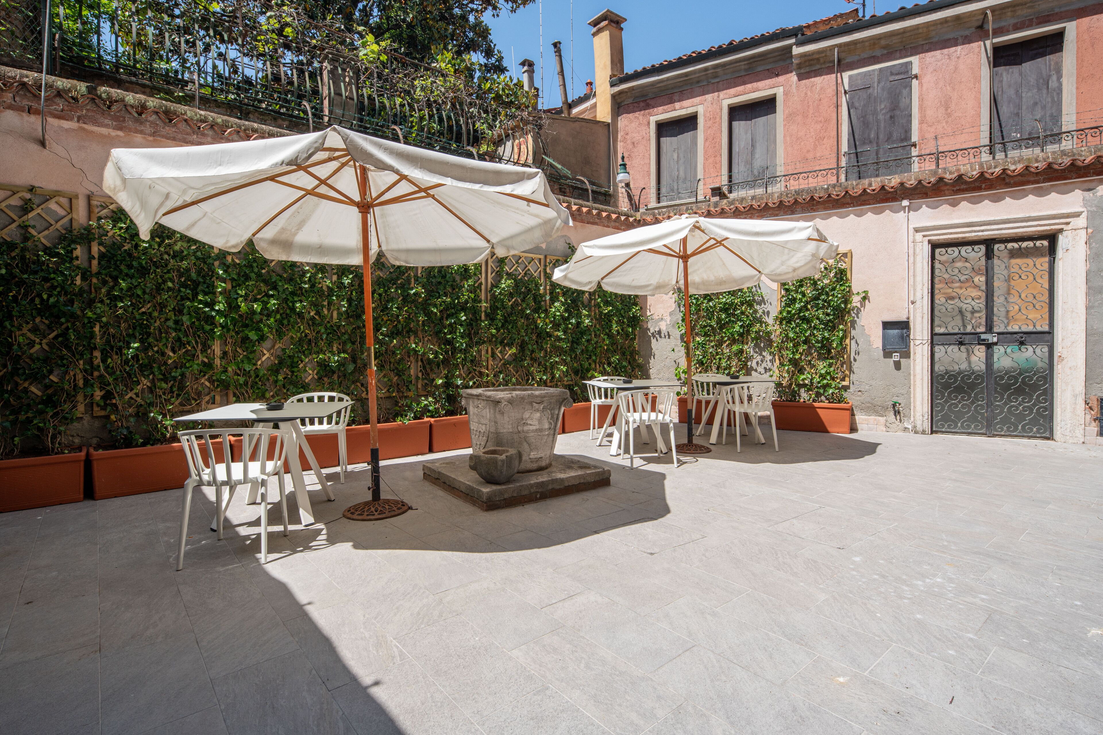 Ein Außenbereich mit einer Terrasse, weißen Tischen und Stühlen, großen Sonnenschirmen und Kübelpflanzen.