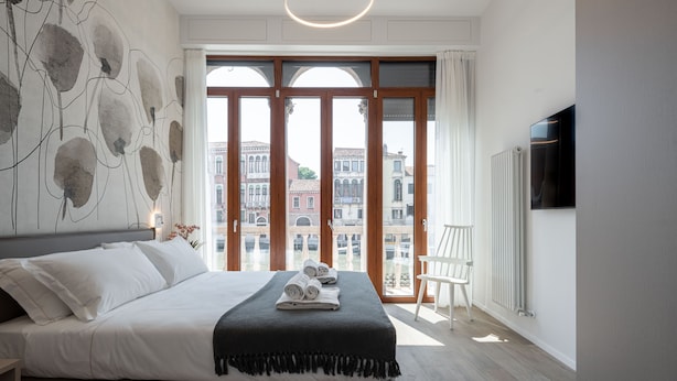 A modern bedroom with a large bed, a view of buildings through large windows, and a radiator on the wall. - Palace Canal View by Wonderful Italy (Venice)