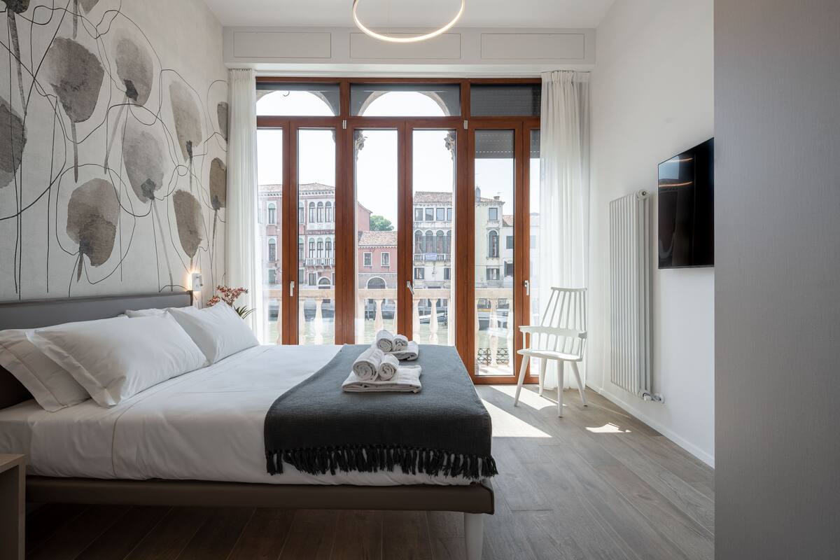 Une chambre moderne avec un grand lit, une vue sur les immeubles à travers de grandes fenêtres et un radiateur au mur.
