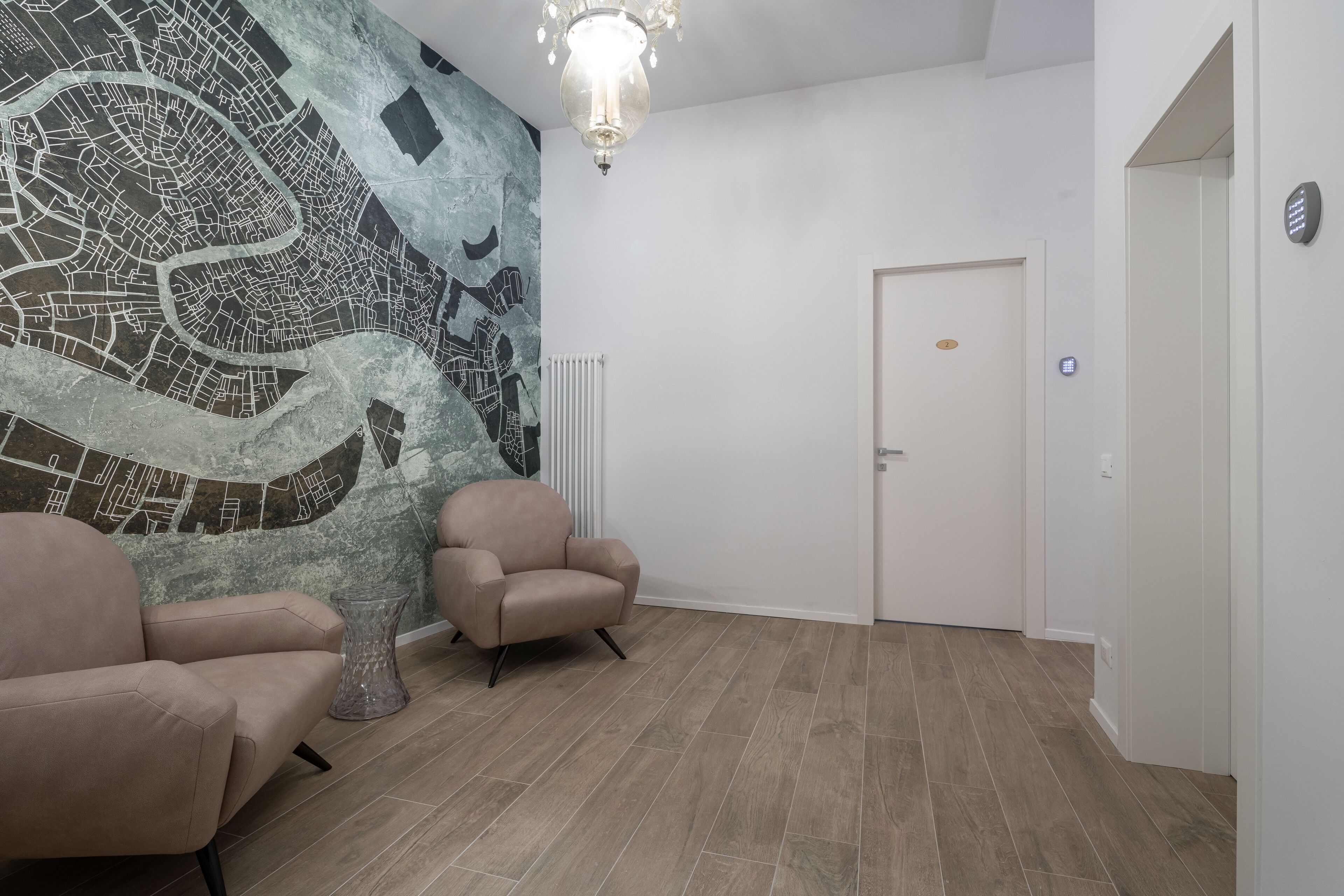 Een kamer met een stadsgezicht muurschildering, twee fauteuils, een kroonluchter en een deur.