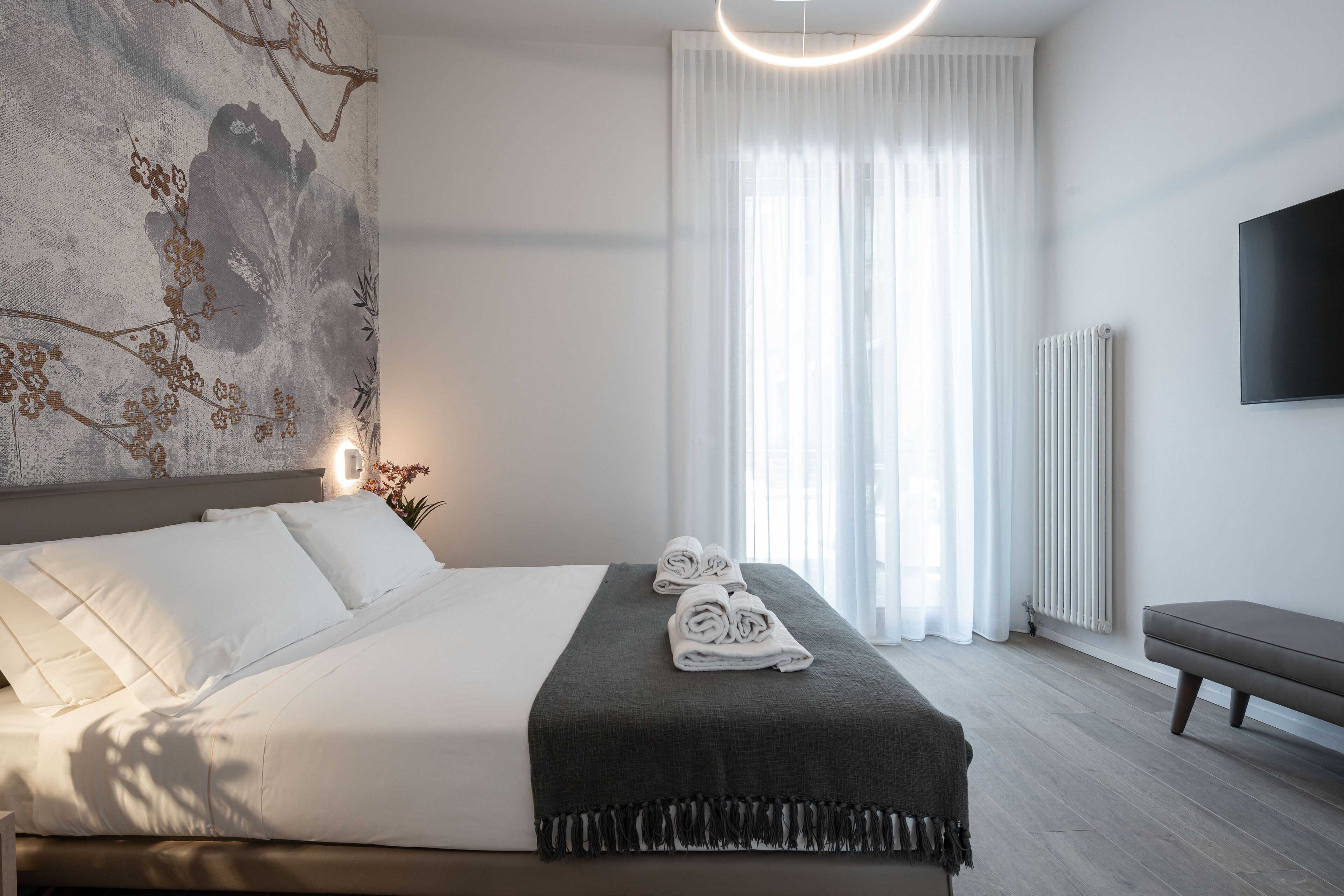 A modern hotel room with a large bed, a bedside table, a flat-screen TV, and a large window with sheer curtains.