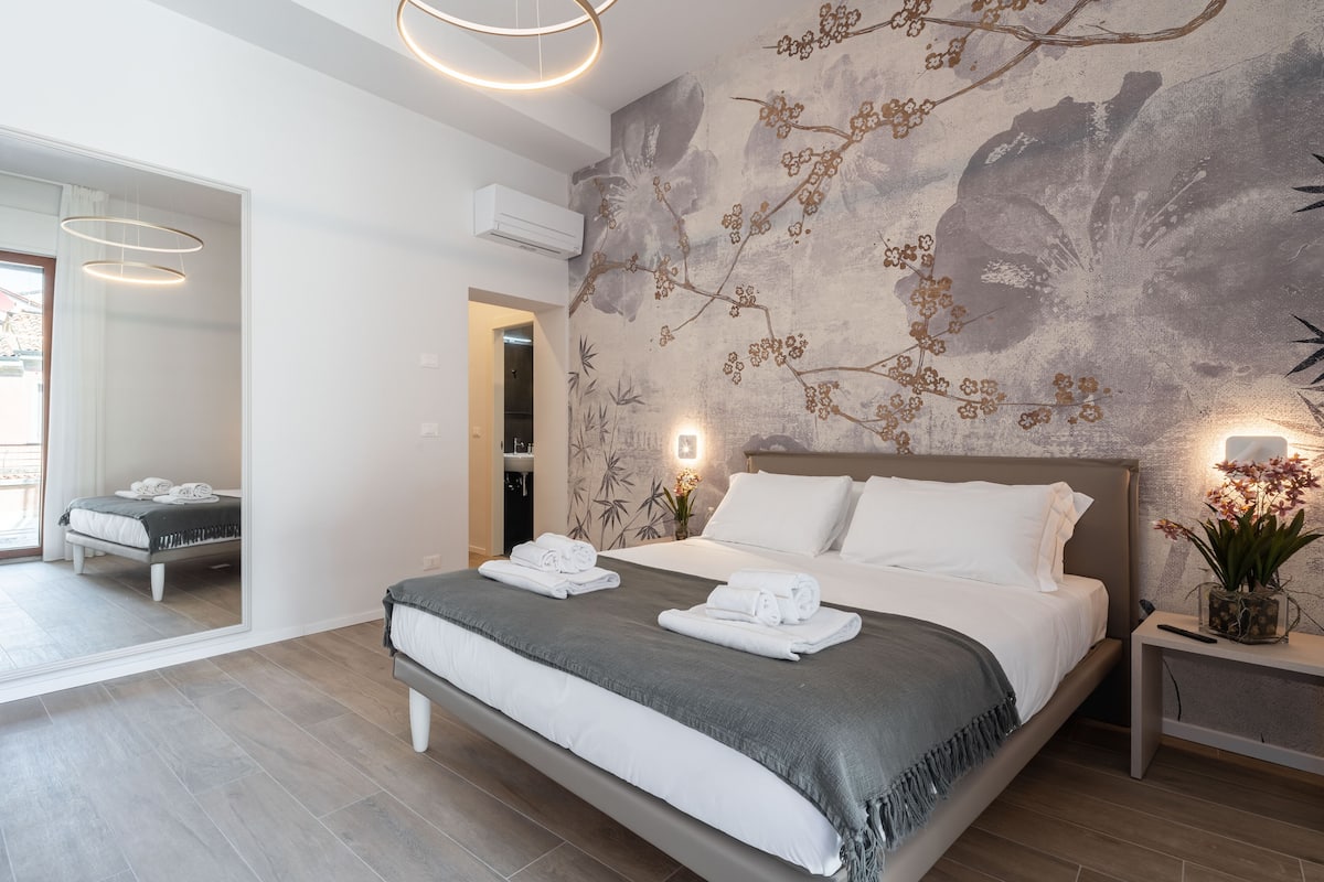 Une chambre moderne avec un grand lit, un climatiseur fixé au mur et une table de chevet agrémentée d’une lampe et d’un vase de fleurs.