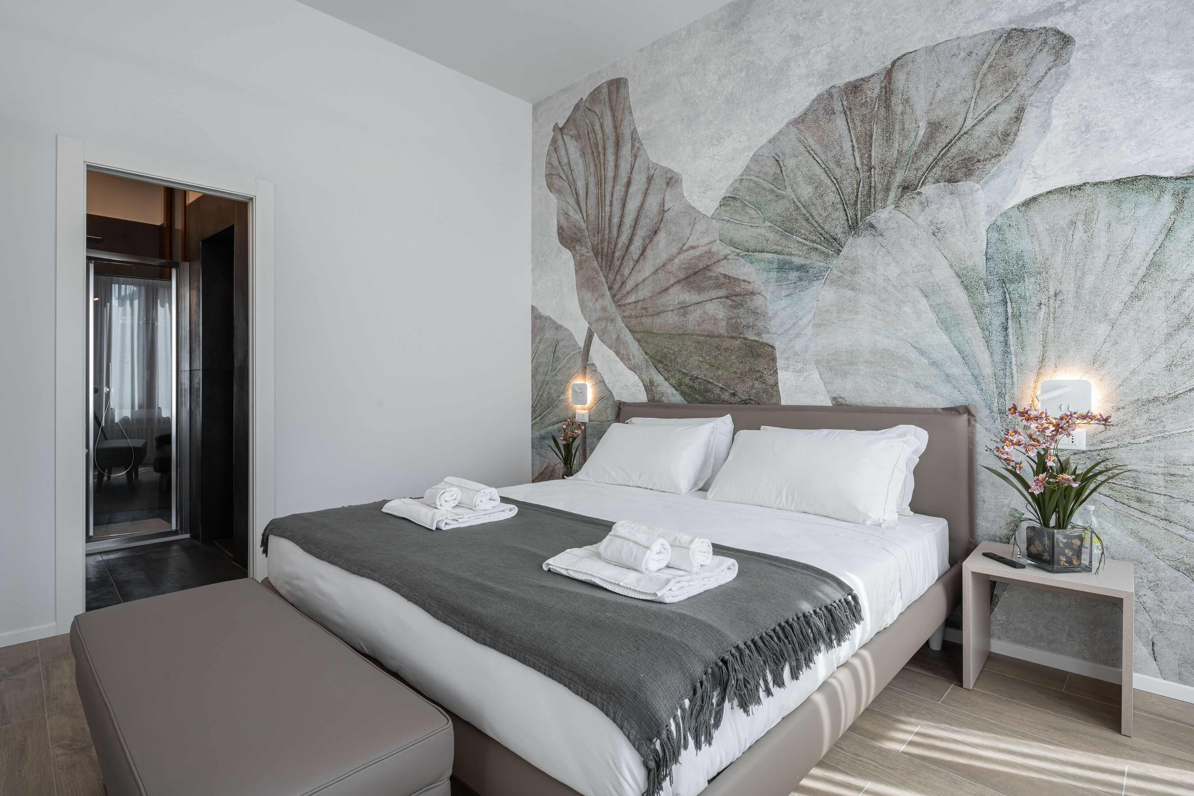 A modern bedroom with a large bed, a bench, a nightstand with a plant, and a wall with a leaf mural.