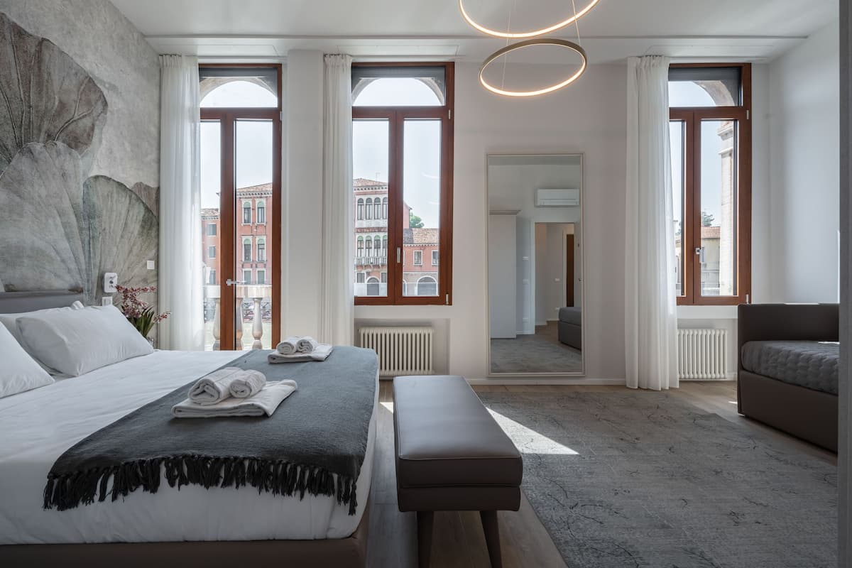 Une chambre moderne avec un grand lit, un banc et une vue sur les immeubles à travers de grandes fenêtres.