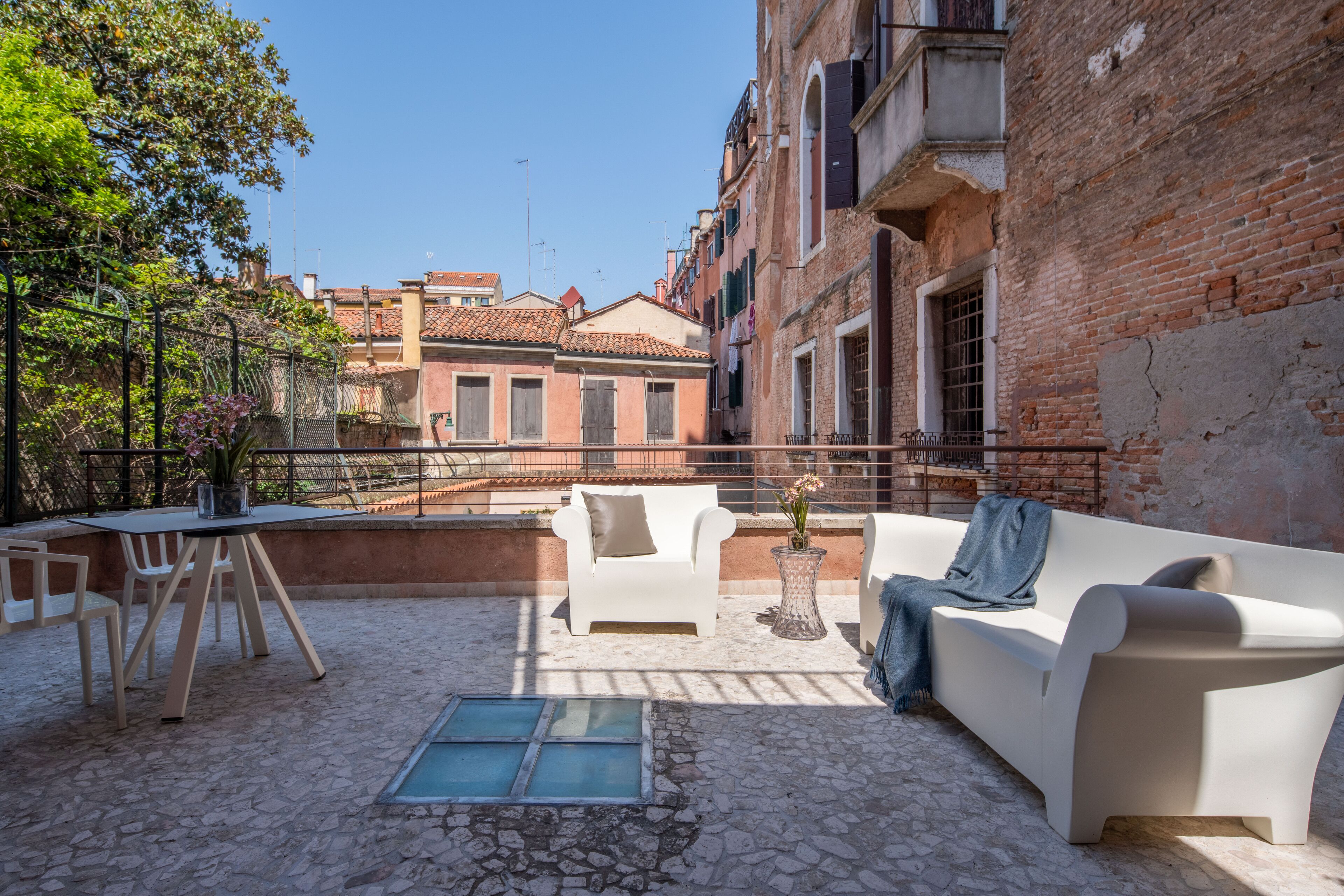 A courtyard with a white sofa, a small table, and chairs. There is a brick wall on one side and a metal fence on the other.